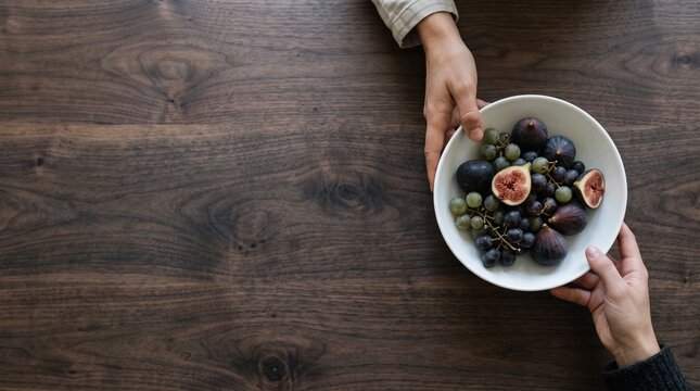 Overhead view of hands holding a bowl of fresh figs and grapes on a dark wooden table