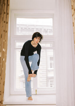 Woman Sits on a Windowsill and Adjusts Jeans in a Bright Room