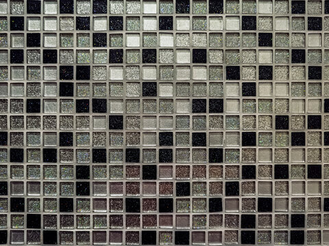 View of decorative mosaic tile wall featuring geometric grid pattern with glossy black, shimmering silver, and white small square tiles as abstract background. Mosaic tile wall texture and background.