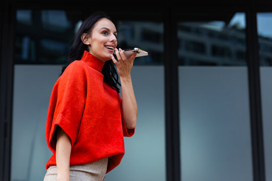 Woman in red sweater smiling, recording a voice message on smartphone, communicating easily with modern technology in a dynamic urban environment outdoors