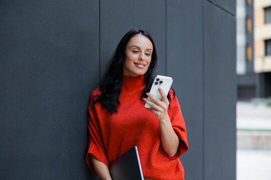 Professional woman smiling, standing outside an urban building, holding a smartphone and a laptop, engaging in digital communication, working remotely, or enjoying a modern connected lifestyle
