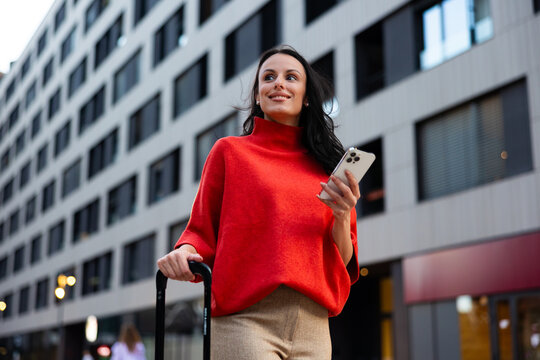 Young businesswoman wearing red sweater and pulling luggage on a city street, looking away, holding smartphone, traveling for work in an urban environment