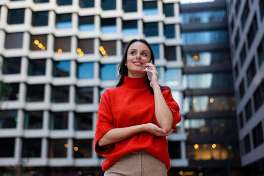 Woman in red sweater making a call on smartphone, looking up and smiling in an urban setting, communicating and connecting in a modern city environment