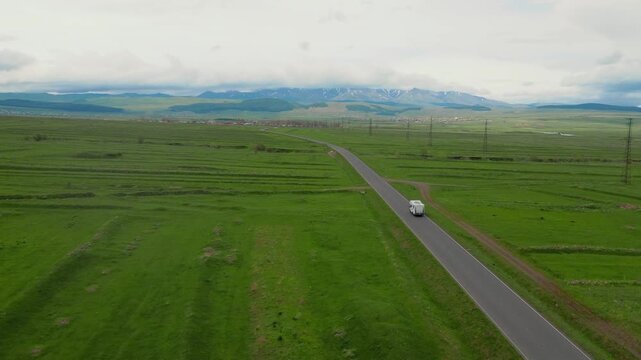 A white camper rides on an empty asphalt road through a vast green landscape with snow-capped mountains in the background, embodying the concept of freedom, travel and adventure vacation in Georgia