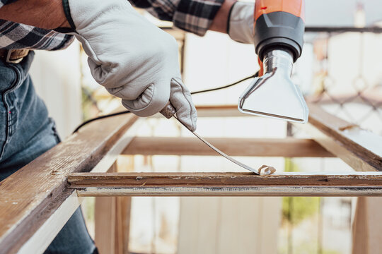 Adult carpenter wearing protective leather gloves, using a heat gun and a wood scraper to remove paint from an old wooden window. Construction industry, carpentry. Restoration. 