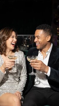 laughing diverse couple in elegant evening clothes clink champagne coupes while seated on green velvet sofa. joyful romantic celebration, nightlife and shared moments at festive party.