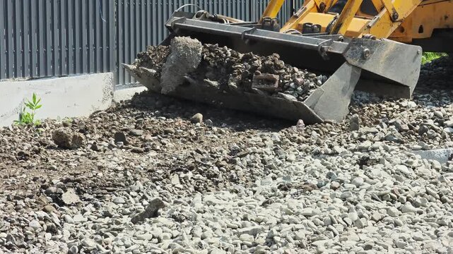 Close up of a yellow backhoe loader scooping soil, rocks, and debris from a gravel surface during road construction, moving the rubble to clear the ground for new development and earthworks