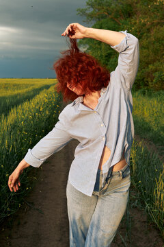 A red-haired woman poses playfully on a rural path beside green crops.