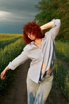 A red-haired woman poses playfully on a rural path beside green crops.
