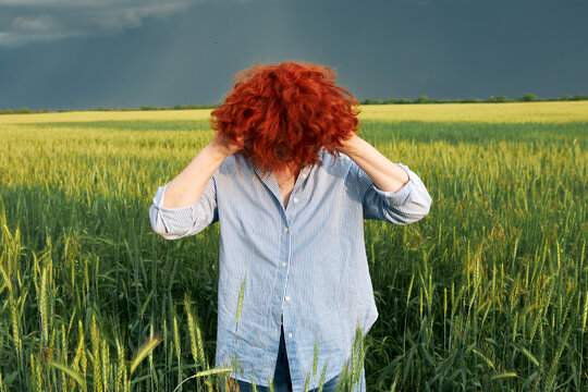 Red-haired woman adjusting hair in green grain field beneath dramatic cloudy skies