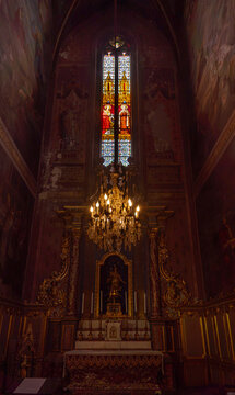 A vertical, low-angle view of the interior of a Neo-Gothic chapel, featuring a gilded altarpiece, frescoes on the walls, a stained-glass window 