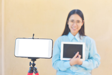 Young woman using mobile device on tripod to create content while holding a tablet, capturing modern technology and creative lifestyle in a bright setting © Suriyo