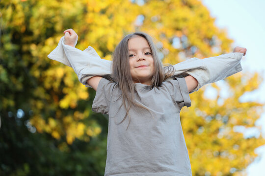 Smiling Young Girl Holding Sweater Over Shoulders With Arms Raised In Autumn Park, Standing Against Soft Golden Foliage With Low Angle View And Natural Daylight Portrait Background