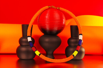 ibrant African beaded bracelet featuring bold orange, yellow, red, blue and white patterns, styled with sculptural vases against a striking red and orange backdrop for a colourful fashion still life. © Valerie