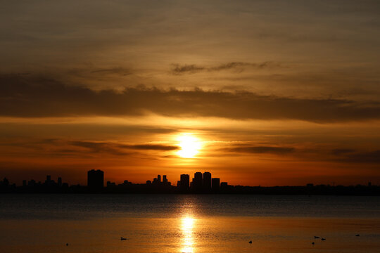 Dramatic orange sunset over a silhouetted city skyline with glowing reflections across calm water and birds in the foreground.