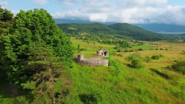 Happy young friends waving and rejoicing on top of a hill as the drone flies away, revealing a beautiful green valley with mountains in Georgia's Racha, a lake and a village road on a sunny day with c