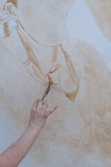 Vertical close-up shot of an artist's hand using a paintbrush to follow pencil sketch lines on a large wall mural. © Phoscar