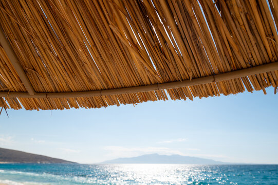 Thatched beach umbrella frames a view of the sea and distant coastline in daylight