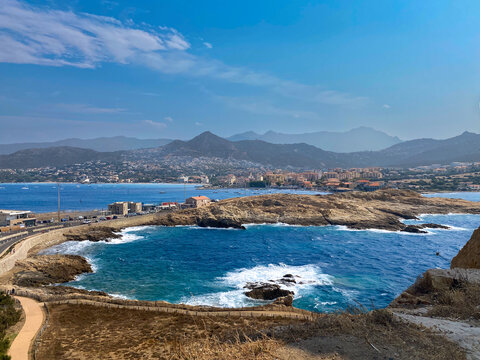 Landscape of &Icirc;le de la Pietra in L'&Icirc;le-Rousse, Corsica, France. blue Mediterranean Sea with crashing waves, connecting causeway, and town nestled against mountains under a blue sky.