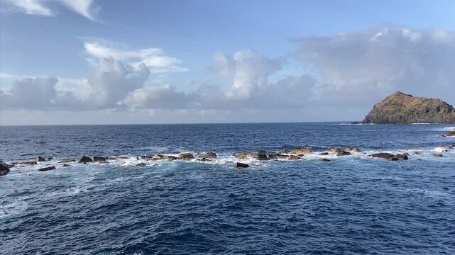 Ocean waves break over dark rocks beside a distant rocky island Roque de Garachico against blue cloudy sky in Tenerife, Canary Islands, Spain,4K.