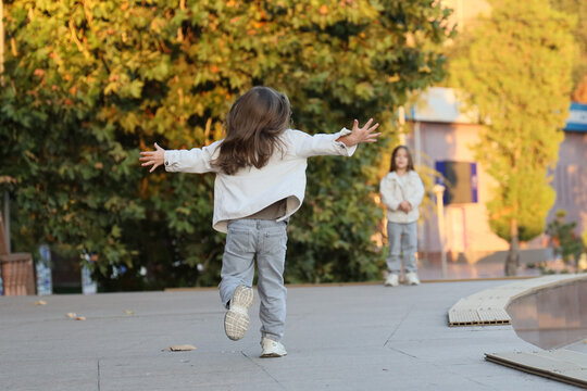 Child Running With Arms Wide Open On Outdoor Walkway In Warm Golden Autumn Light, Another Child Standing Nearby In Soft Focus, Playful Family Mood And Spacious Background Nature Setting Outdoors.