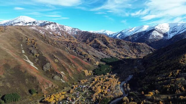 Aerial drone footage of an autumn mountain landscape with rolling brown hills, golden yellow trees, and dense evergreen forest patches. Snow-covered mountain peaks rise in the background beneath a bri