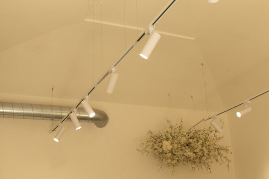 white cylindrical track spotlights on metal rail illuminate beige high ceiling in modern interior, with visible wires, ventilation duct, and ceiling flowers