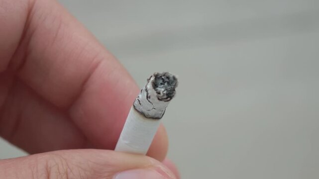 Close up view of a person's hand holding a lit cigarette, showing the ember glow and the ash slowly forming and growing at the tip, illustrating addiction and unhealthy habits