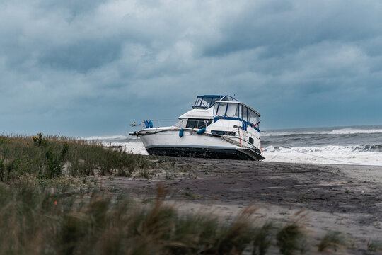 A cabin boat rests on the beach along the New York coast. A white motorboat with blue trim sits on dark sand near breaking waves beneath gray clouds.