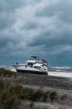 A boat rests beneath heavy clouds on the New York coast. A motorboat rests above the surf with dune grass in the foreground and a broad expanse of sky.