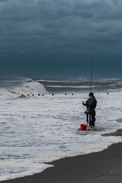 An angler stands among waves in a vertical view of the New York coast. A person stands in foamy water holding a rod beside a red bucket beneath layered clouds.