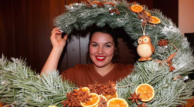    Candid portrait of a woman playfully holding a large winter wreath with dried oranges and a wooden owl. Dark wood background. Joyful holiday spirit.
