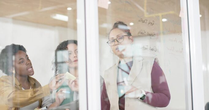 Female team passing sticky notes leading bespectacled woman in vest writing on glass and planning