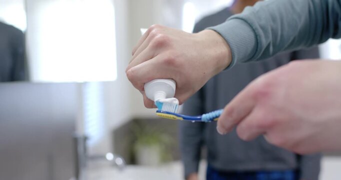 Foreground hands are squeezing paste onto blue brush, man is brushing at sink for morning care