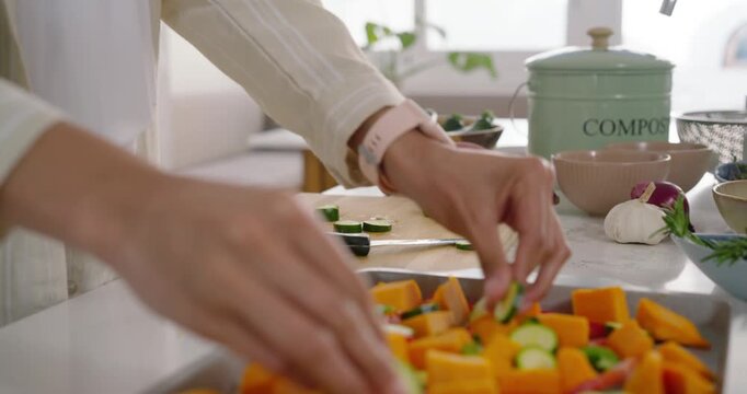 Home cook is reaching with knife, slicing zucchini and arranging veg on tray for roasting
