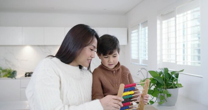 Asian mother leaning and speaking softly, guiding child son moving abacus beads for math in kitchen