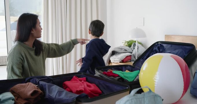 Asian mother and child packing luggage on bed, mother passing sweater and guiding folding for trip