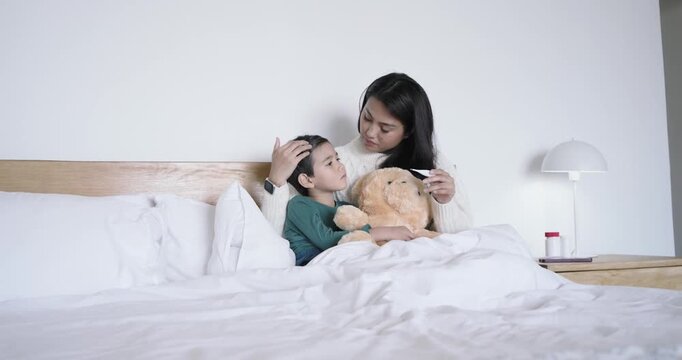 Asian mother checking child's temperature, prompted by fever concern, comforting in bed with teddy