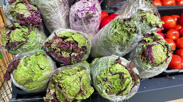 Heads of green and purple napa cabbage wrapped in plastic beside red tomatoes at a grocery store, vegetable display, fresh produce, food shopping