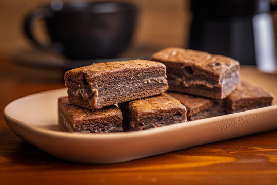 Stacked Homemade Chocolate Brownies on a Plate with Coffee Cup Close-Up in Cozy Cafe Setting