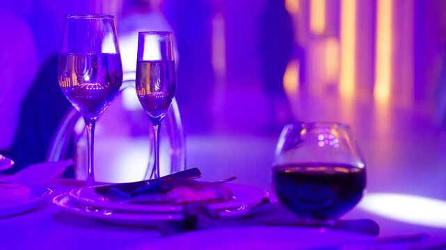 Magenta lighting over wine glasses and dinner plate. Guests dancing at lively evening reception