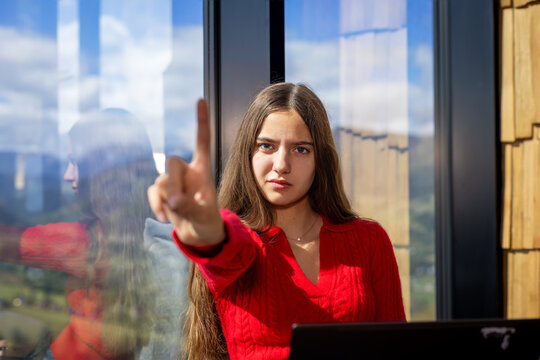 Young woman pointing finger toward camera with serious expression beside laptop, confident gesture in modern interior with panoramic mountain view.