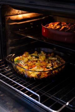 A vegetable casserole bakes on a lower wire rack in an open oven alongside banana bread on the upper shelf. The home-baked dishes highlight culinary preparation and cooking.