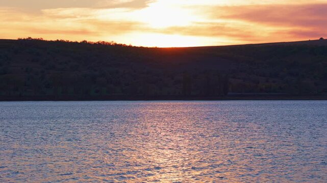 Sunset reflection shimmering on lake waves below hill ridge. Warm evening glow in nature vista