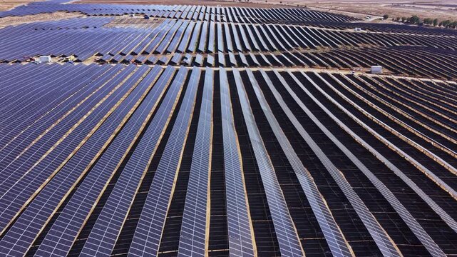 Long rows of solar panels stretch across a solar farm in daylight, viewed from above with the panels arranged in parallel lines.
