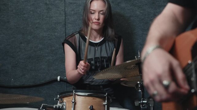 Female drummer playing drums with electric guitarist nearby, focused musician striking cymbals and keeping rhythm during small band rehearsal in music studio.