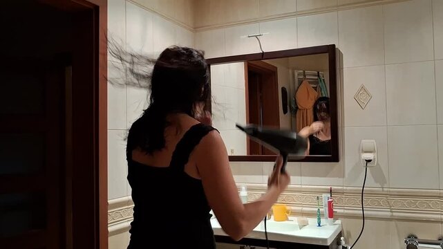 Woman dries her hair with a hair dryer in front of a bathroom mirror