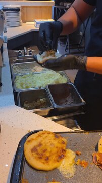 Close-up of a cook wearing black gloves preparing authentic homemade pupusas filled with cheese and beans on a griddle plate during breakfast service