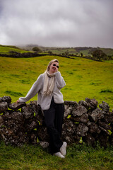 Laughing woman in a grey fleece and scarf leaning on a dry stone wall in green Azorean pastures © Jelena