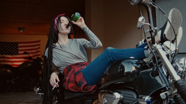 A mechanic woman with dreadlocks leans on a motorcycle in a garage, holding a drink and a cigarette. They wear casual clothes and have long hair with a bandana, with an American flag in the background